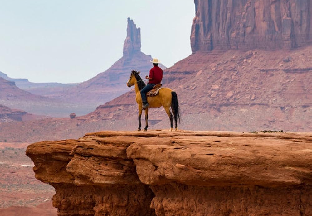 Movie poster for 'The American Frontier: A Film by Edward Frumkin', featuring a cowboy on a horse atop a red rock cliff in a desert canyon landscape, with the title in large tan letters at the top and credits at the bottom.